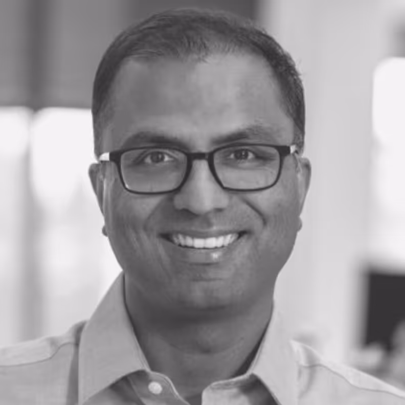 Smiling man wearing glasses and a collared shirt in a softly blurred indoor setting.