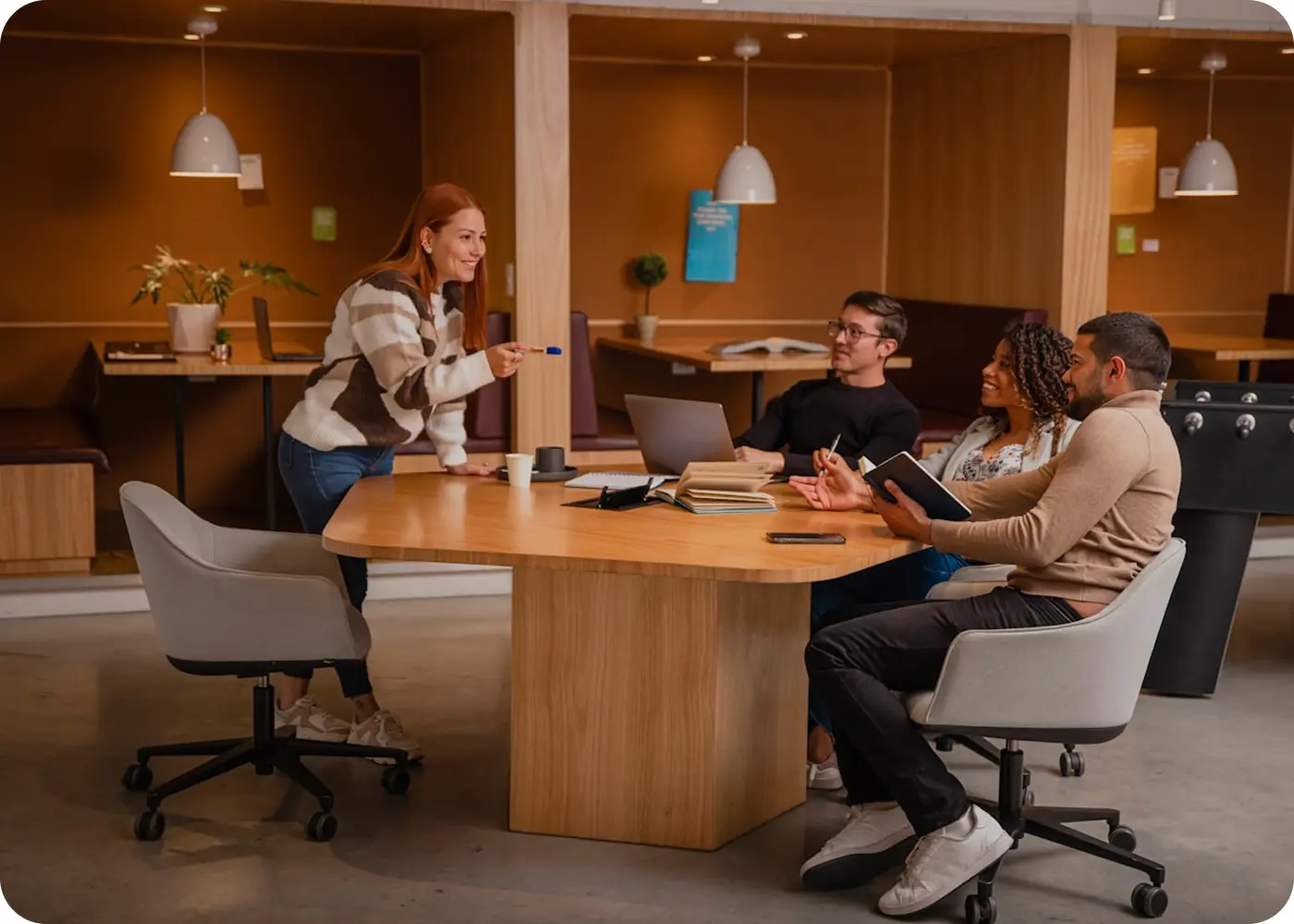 Four colleagues in a modern office having a discussion around a wooden table, with laptops and notebooks.