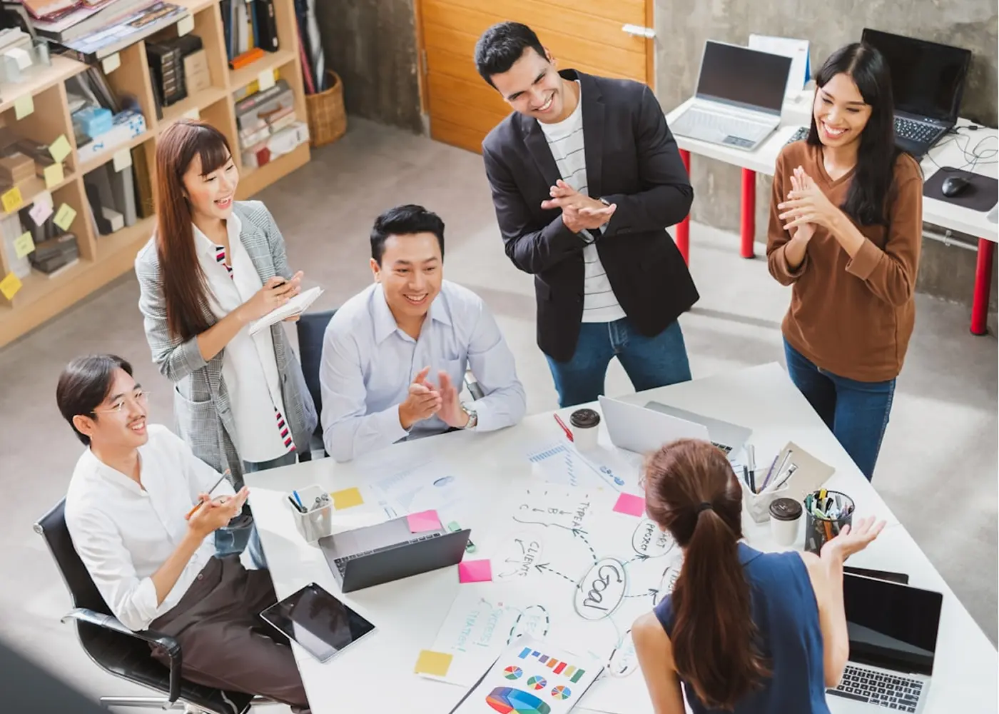 Diverse group of six coworkers smiling and clapping during a meeting around a table with laptops, charts, and notes.
