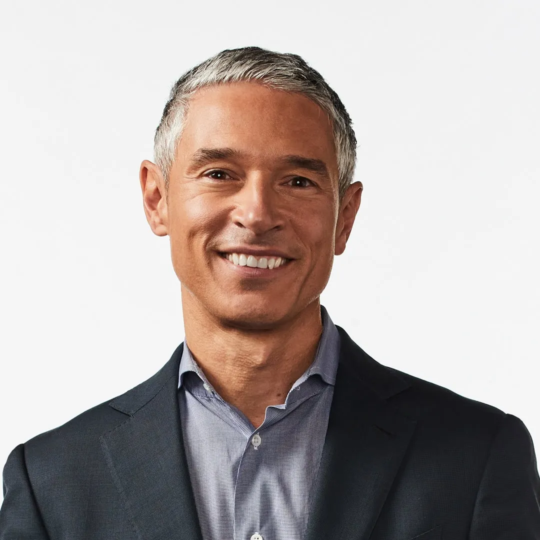 Smiling middle-aged man with gray hair wearing a dark suit jacket and light blue shirt against a white background.
