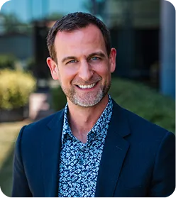 Smiling man with short brown hair and beard wearing a blue blazer and floral shirt outdoors.