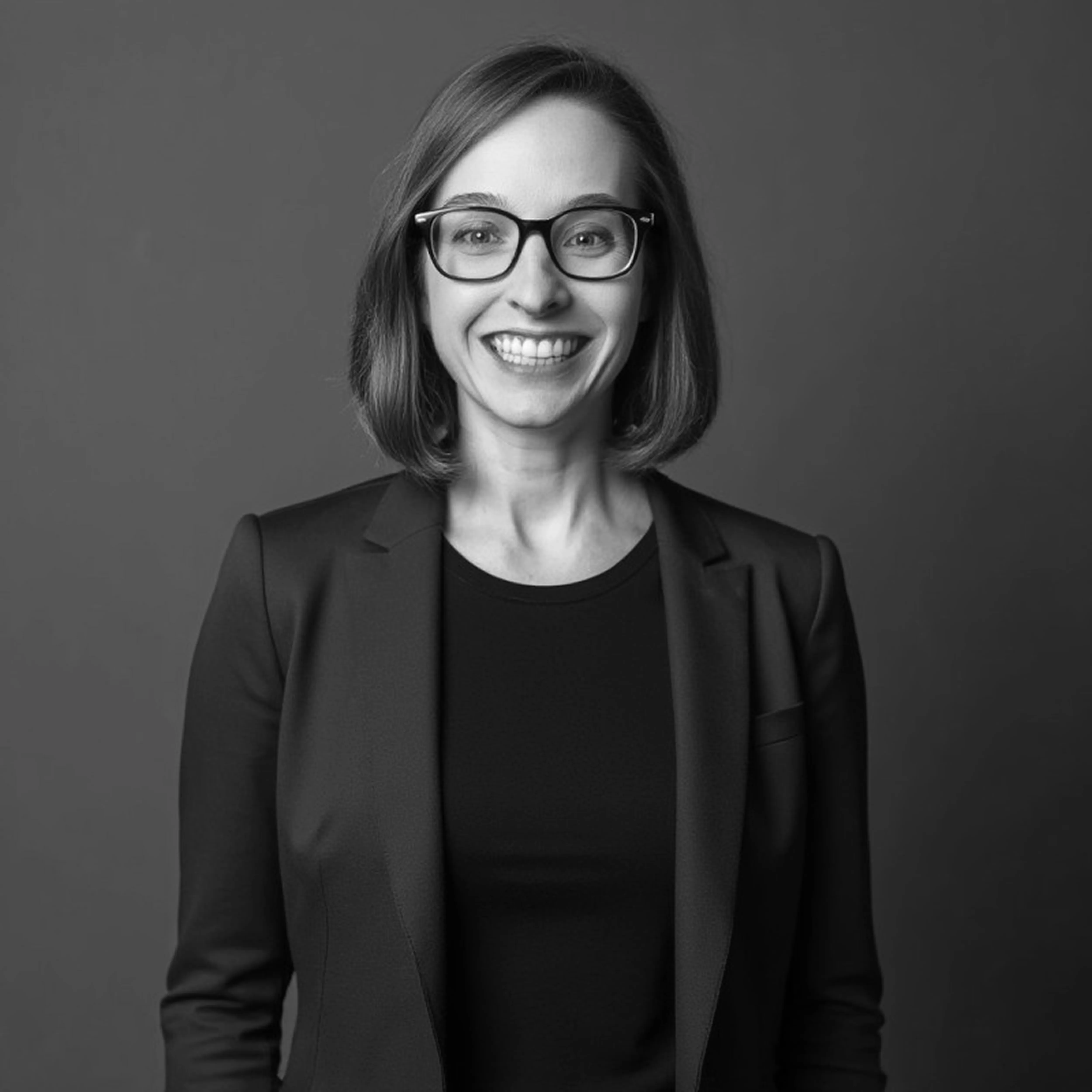 Smiling woman with shoulder-length hair wearing glasses, a black blazer, and a black shirt, against a plain dark background.
