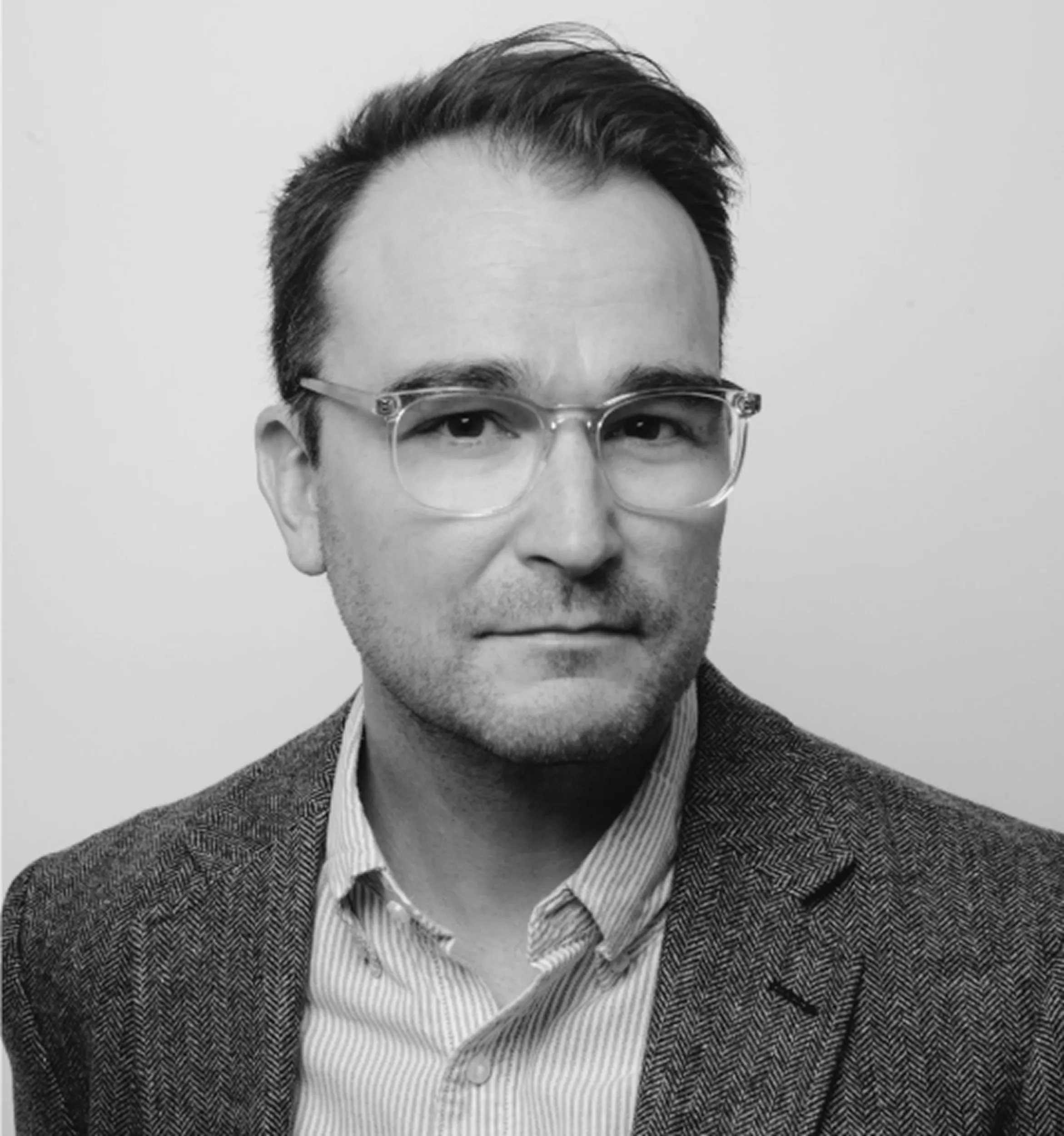 Black and white headshot of a man wearing clear-framed glasses, a striped shirt, and a textured blazer.