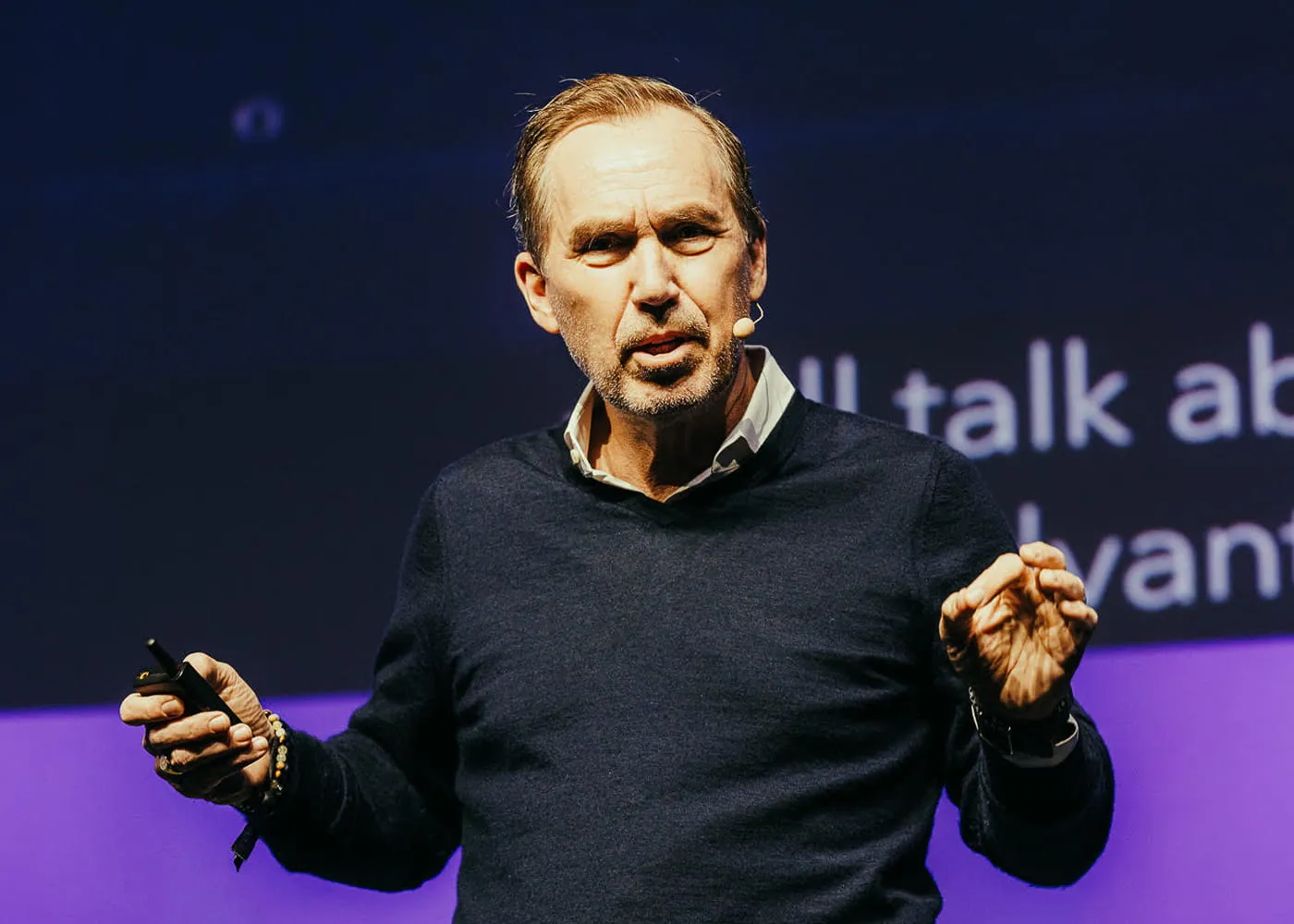 Middle-aged man with a microphone headset speaking on stage holding a clicker, wearing a dark sweater over a white shirt.