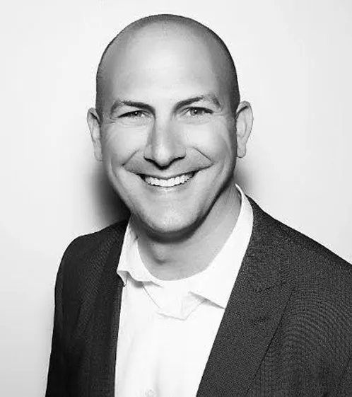 Smiling bald man in a dark suit jacket and white shirt against a plain background.