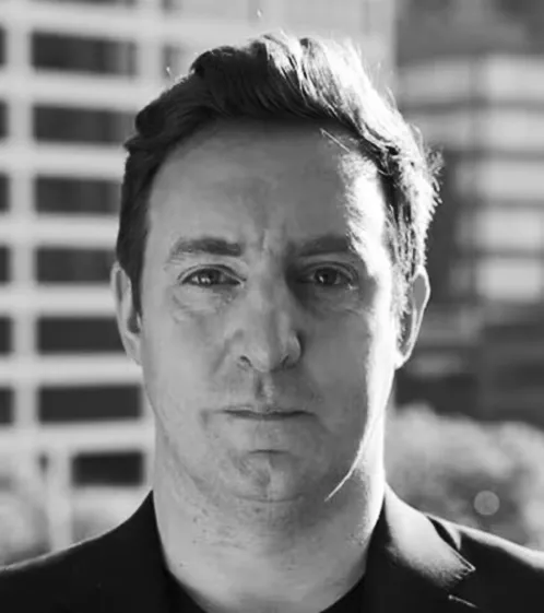 Black and white portrait of a man with short dark hair wearing a dark blazer, standing outdoors with blurred buildings in the background.