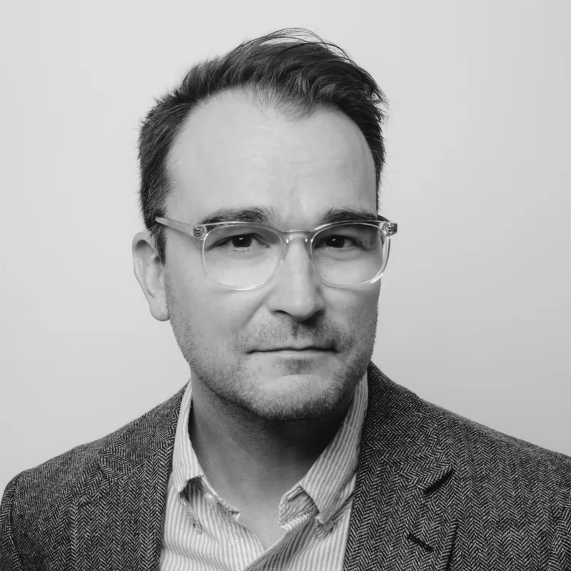 Man wearing clear-framed glasses and a herringbone blazer over a striped shirt, looking at the camera with a neutral expression.