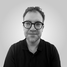 Portrait en noir et blanc d'un homme souriant avec une barbe et une chemise noire à col en V.