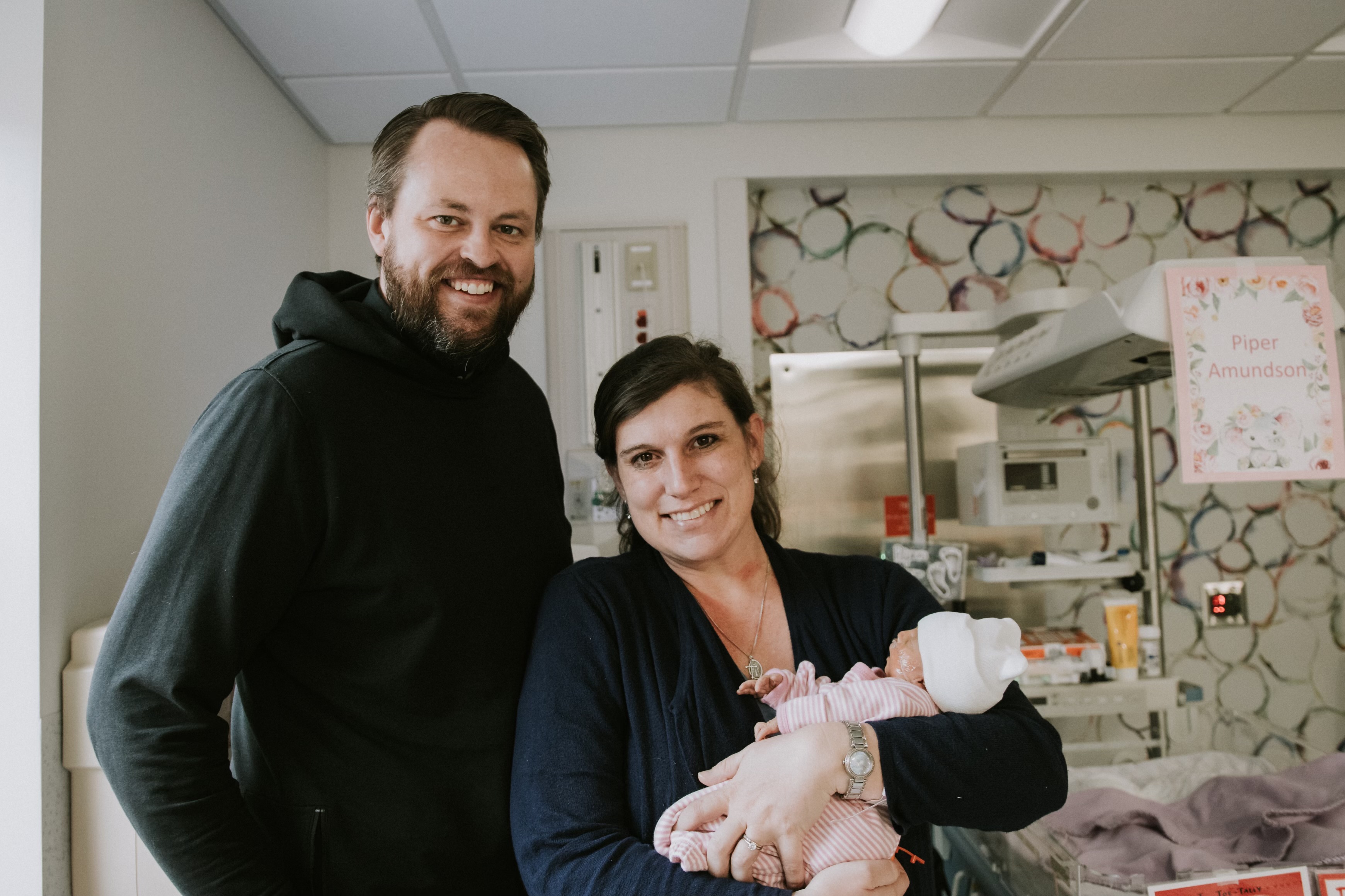 Smiling man and woman holding a newborn baby dressed in pink in a hospital room with a sign that reads Piper Amundson.