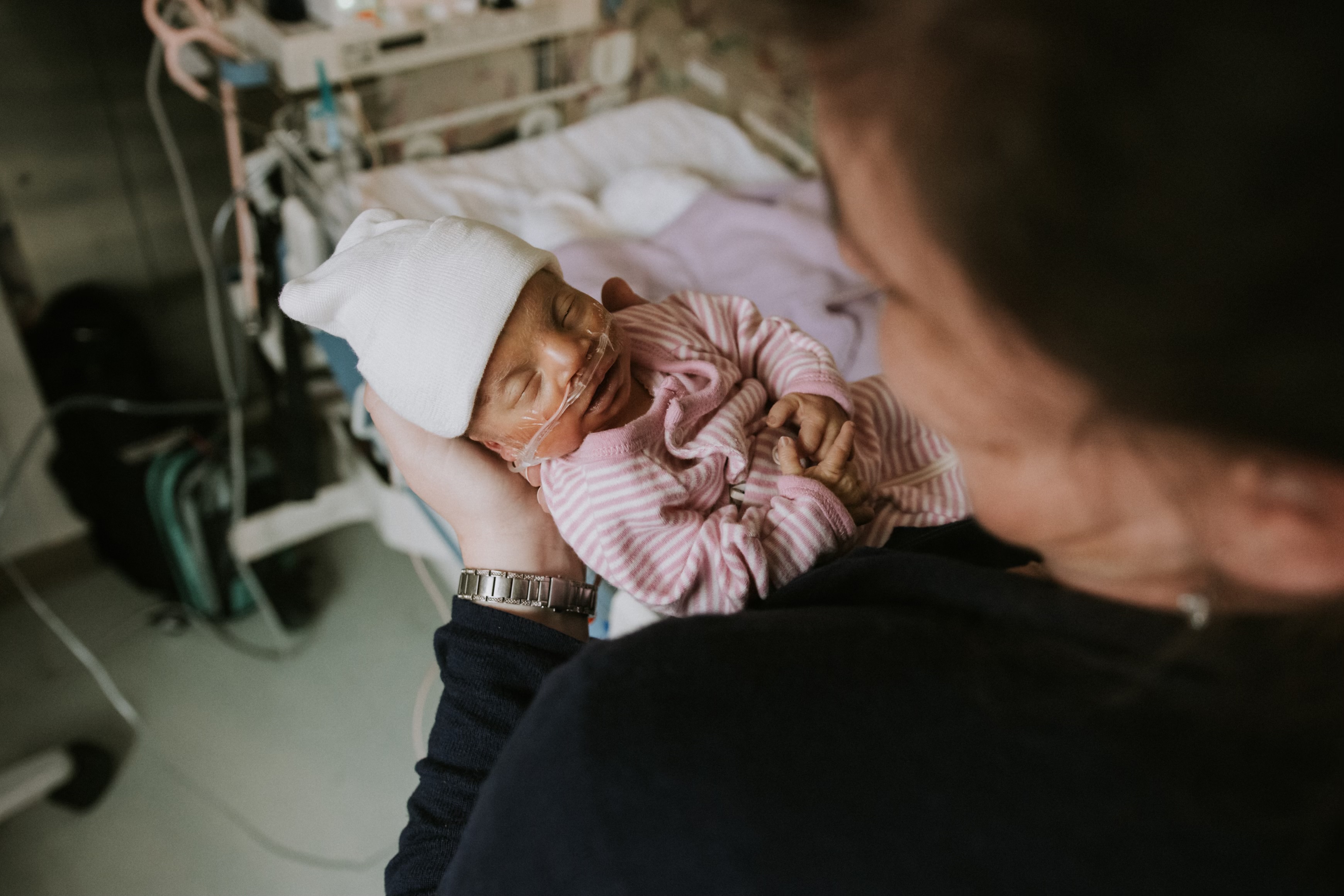 Adult holding a sleeping premature baby wearing a white hat and nasal oxygen tubes in a hospital room.