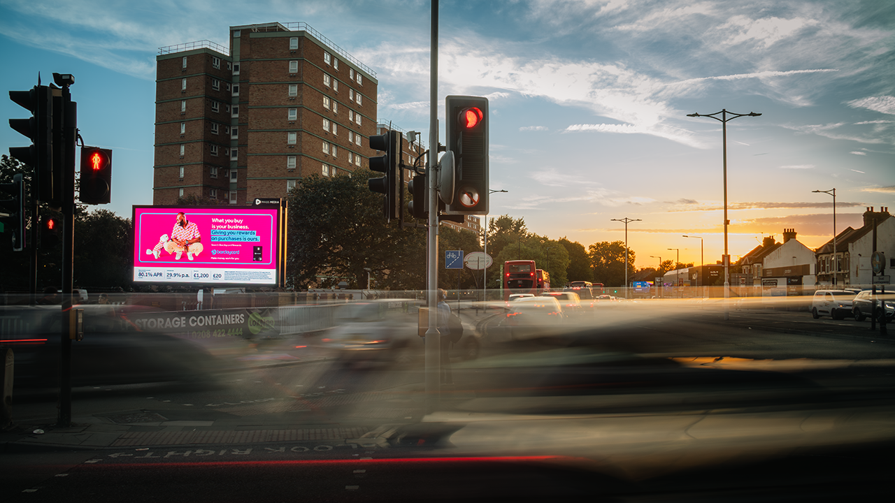Urban street intersection at sunset with red traffic lights, blurred moving vehicles, and a digital billboard on a brown brick building.