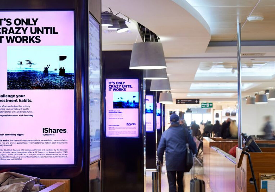 Airport terminal corridor with multiple digital advertisement screens displaying an iShares ad reading 'It’s only crazy until it works', and passengers walking with luggage.