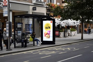 People sitting at a London bus shelter with a digital advertisement for a £4.79 meal deal featuring a sandwich and drink.