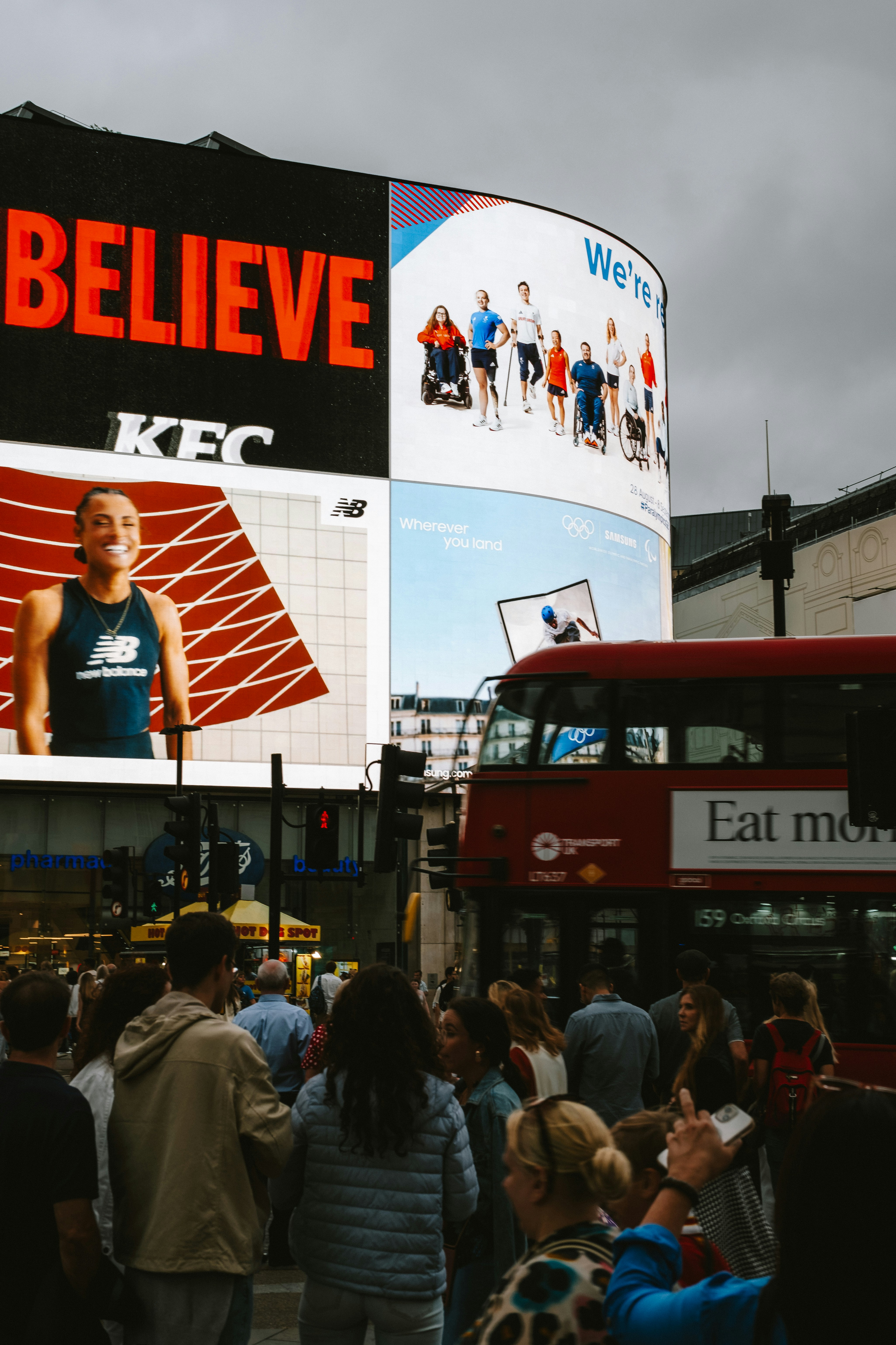 Crowd of people at a busy city intersection with large digital billboards and a red double-decker bus passing by.