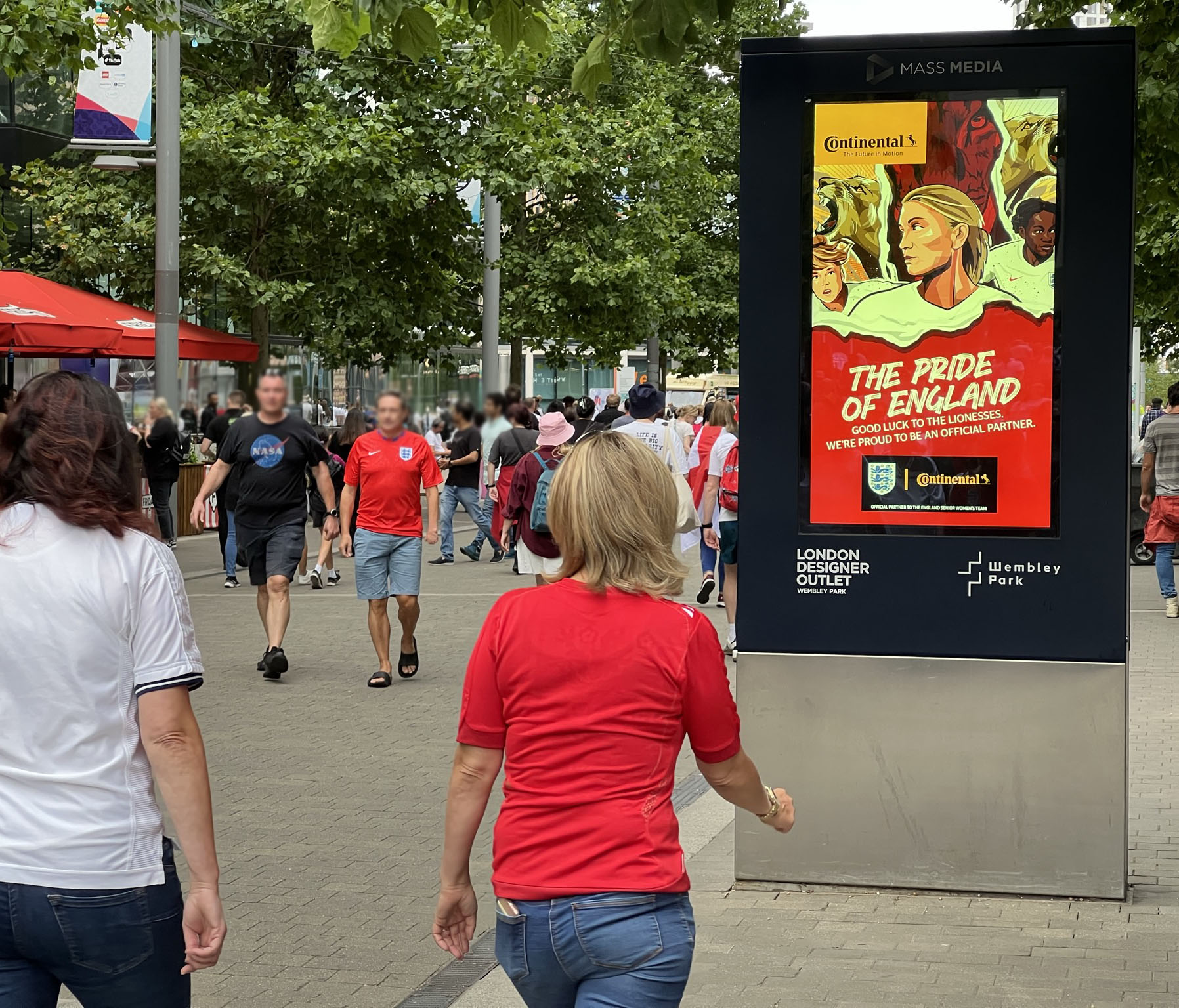 Street scene at Wembley Park with people walking and a digital billboard displaying a 'The Pride of England' advertisement supporting the Lionesses soccer team.