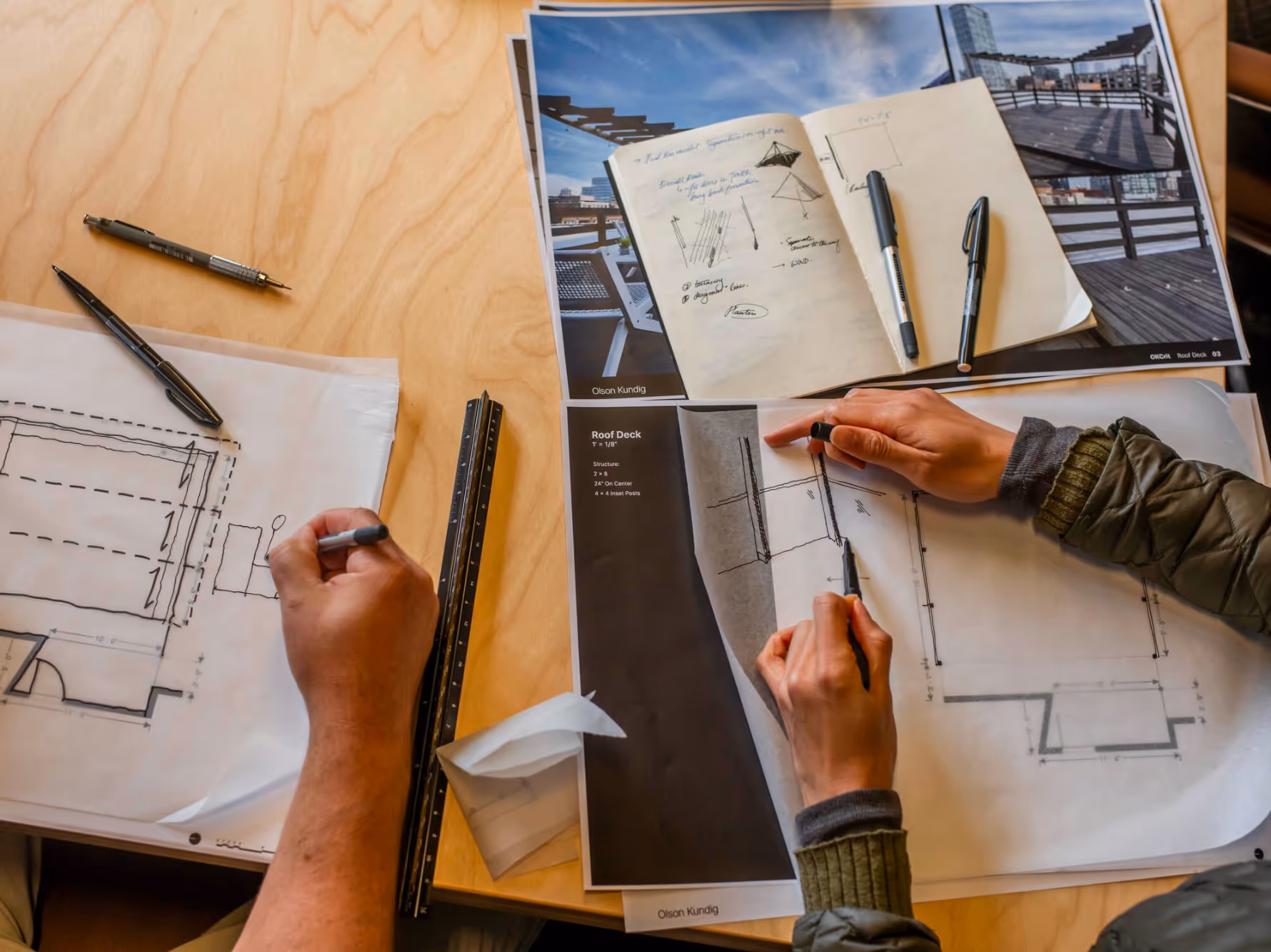 Two people working on architectural blueprints and sketches at a wooden table with pens, ruler, and an open notebook.