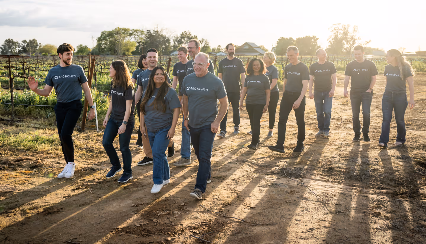 Group of people wearing matching ARO Homes t-shirts walking outdoors on a dirt path with vineyard plants nearby in the evening light.