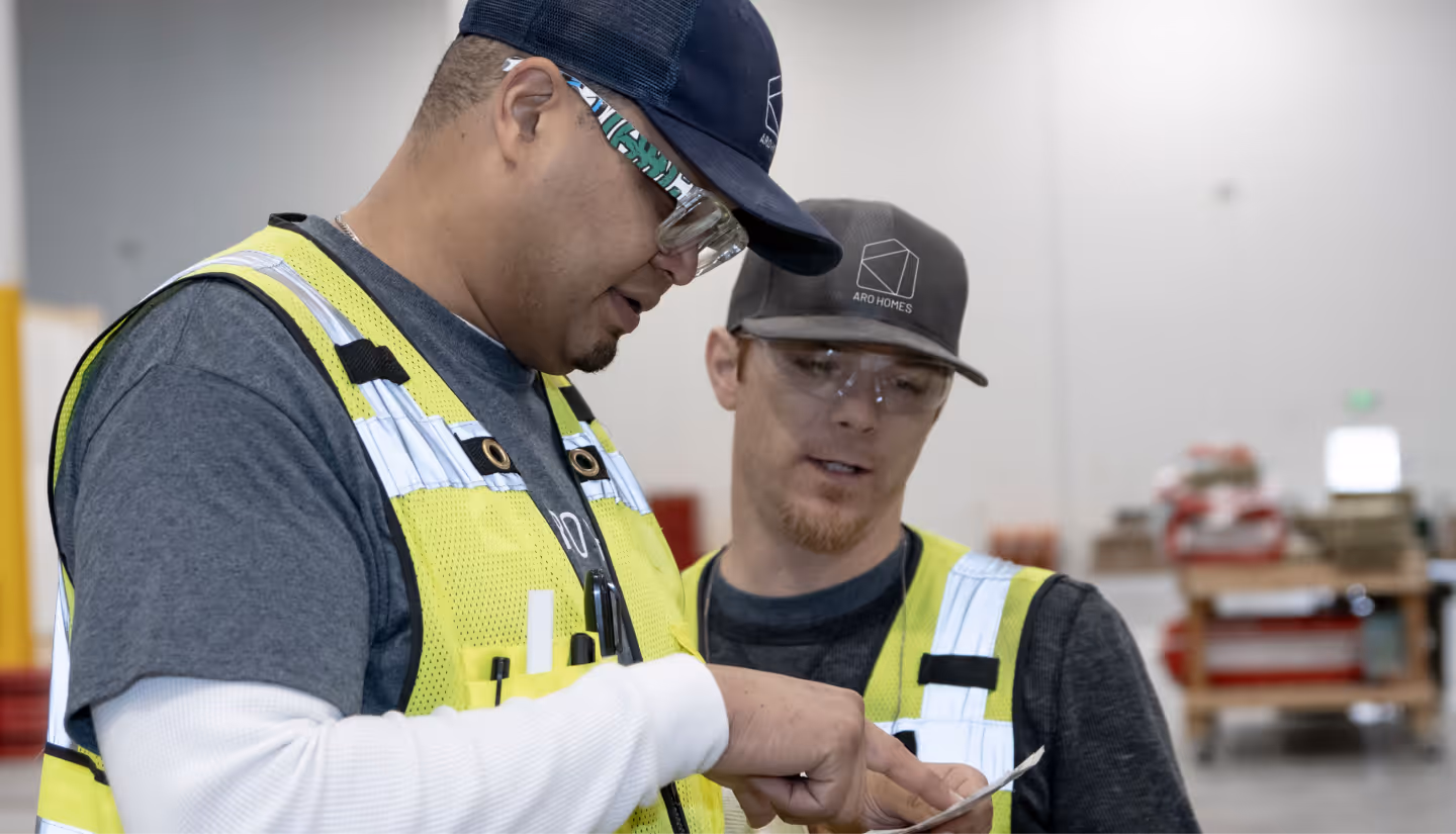 Two construction workers wearing safety vests and glasses reviewing a document inside a warehouse.