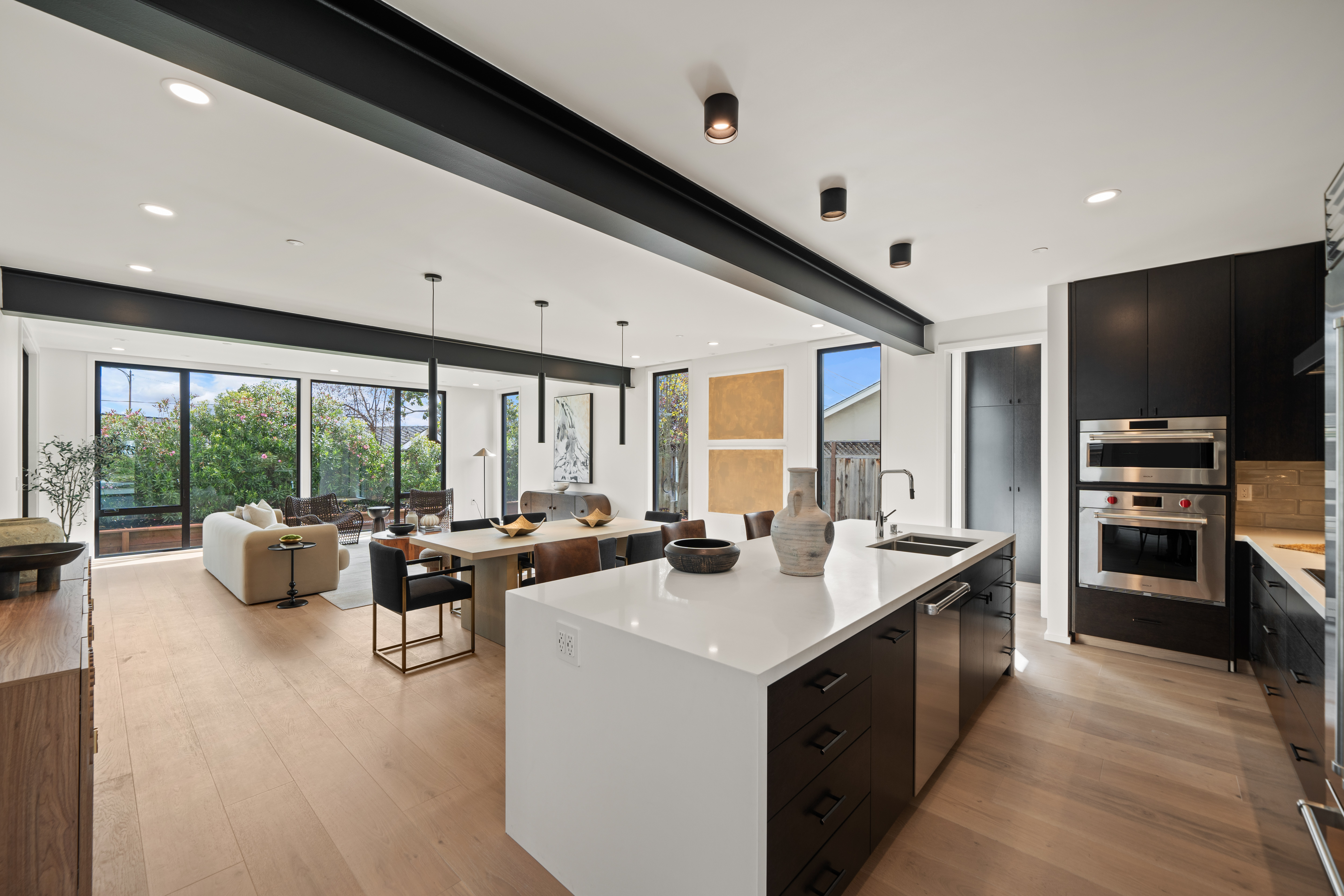 Modern open kitchen and dining area with large island, dark cabinets, wooden floor, and large windows overlooking garden.