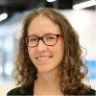 Headshot of a smiling woman with wavy brown hair and glasses, wearing a dark top.