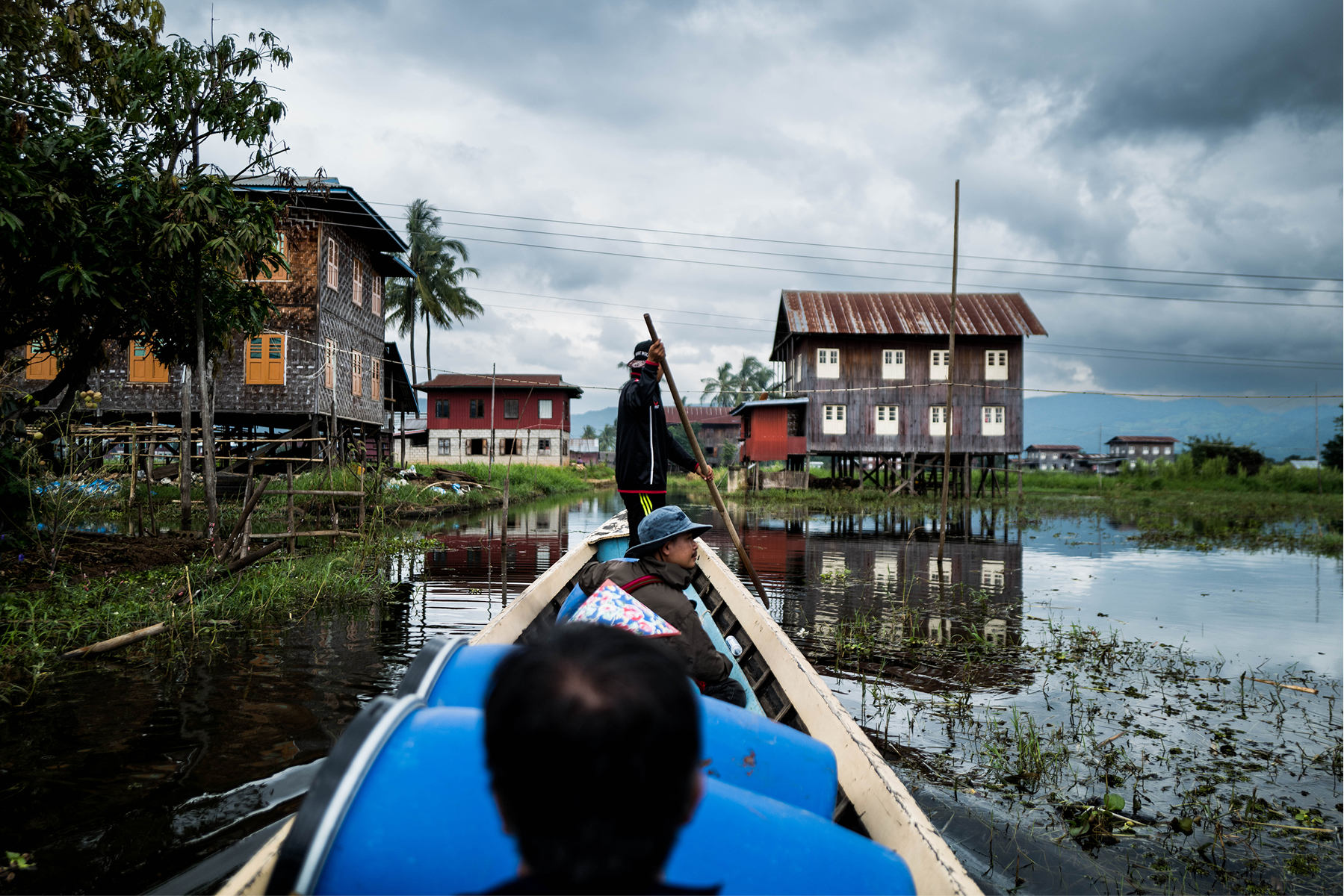 Two people in a wooden boat navigating a waterway with stilt houses on either side under a cloudy sky.