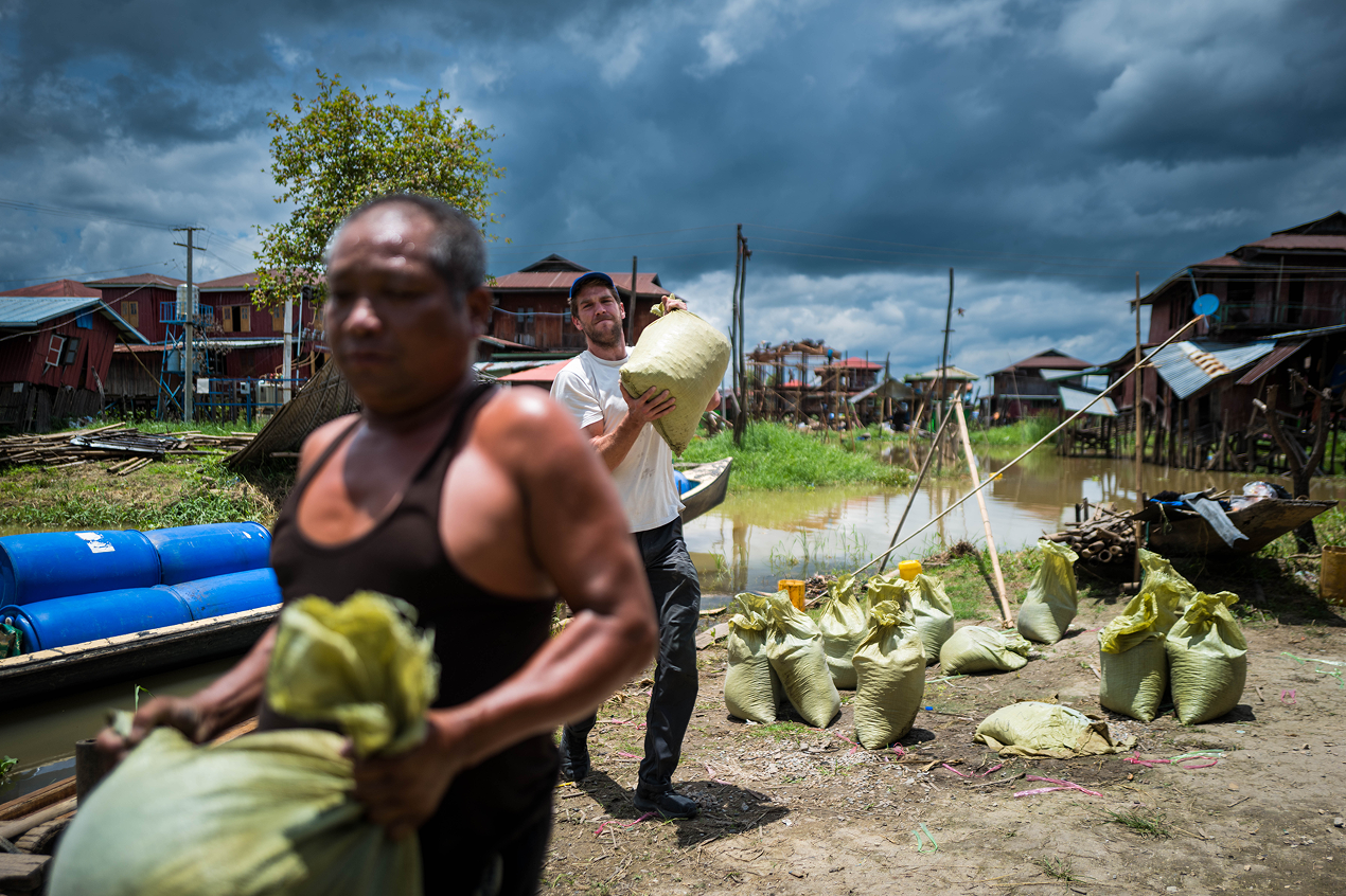 Two men carrying large green sacks near a waterway with wooden houses on stilts under a cloudy sky.