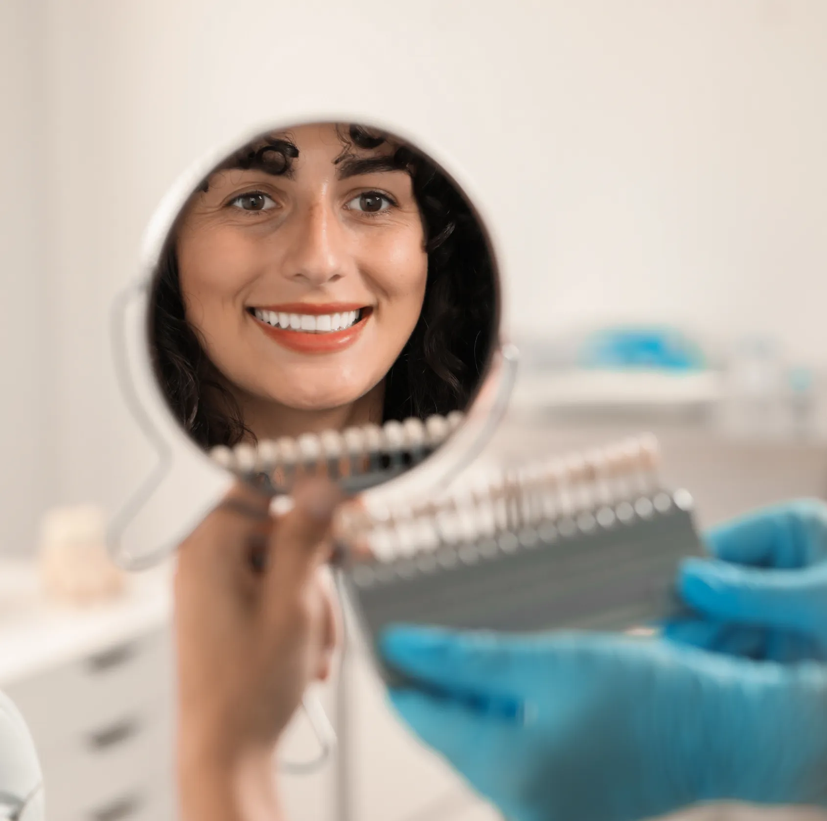 Smiling woman with curly hair looking at her teeth in a round mirror being held by a dental professional wearing blue gloves in a dental office.