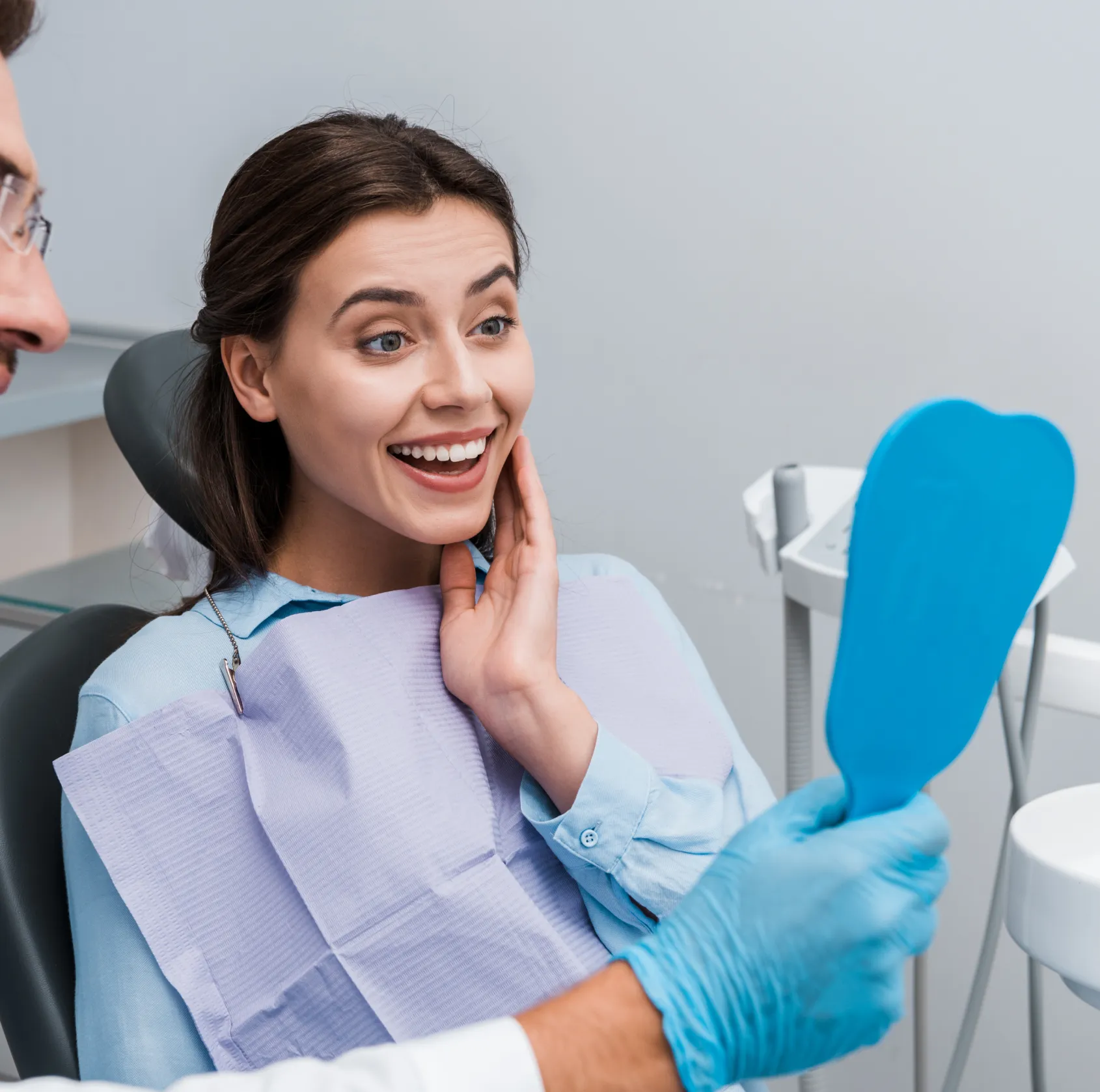 Smiling woman sitting in dental chair looking at her teeth in a blue hand mirror held by a dentist wearing blue gloves.