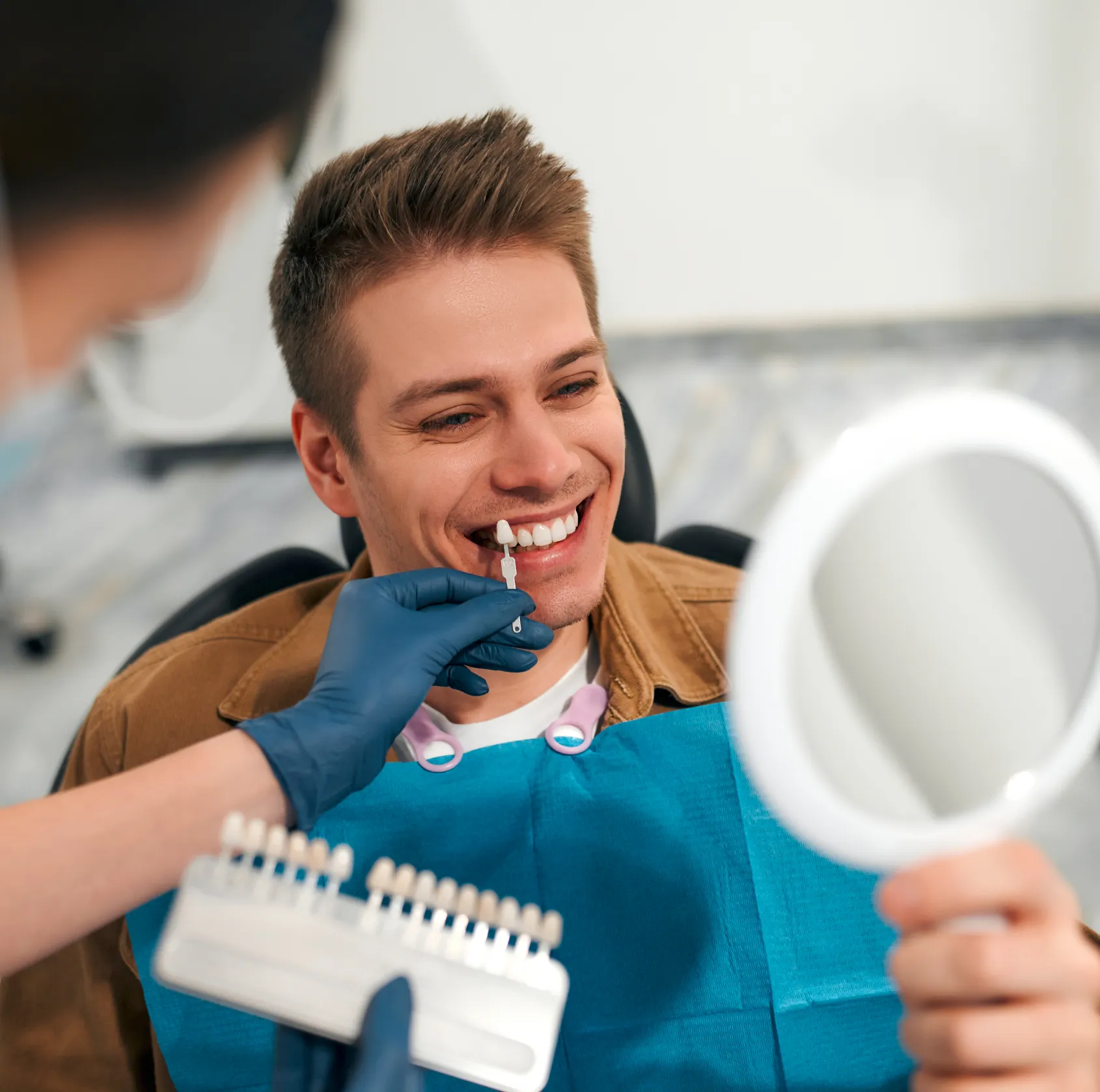Man in dental chair smiling as dentist holds a veneer shade guide up to his teeth and he looks in a handheld mirror.