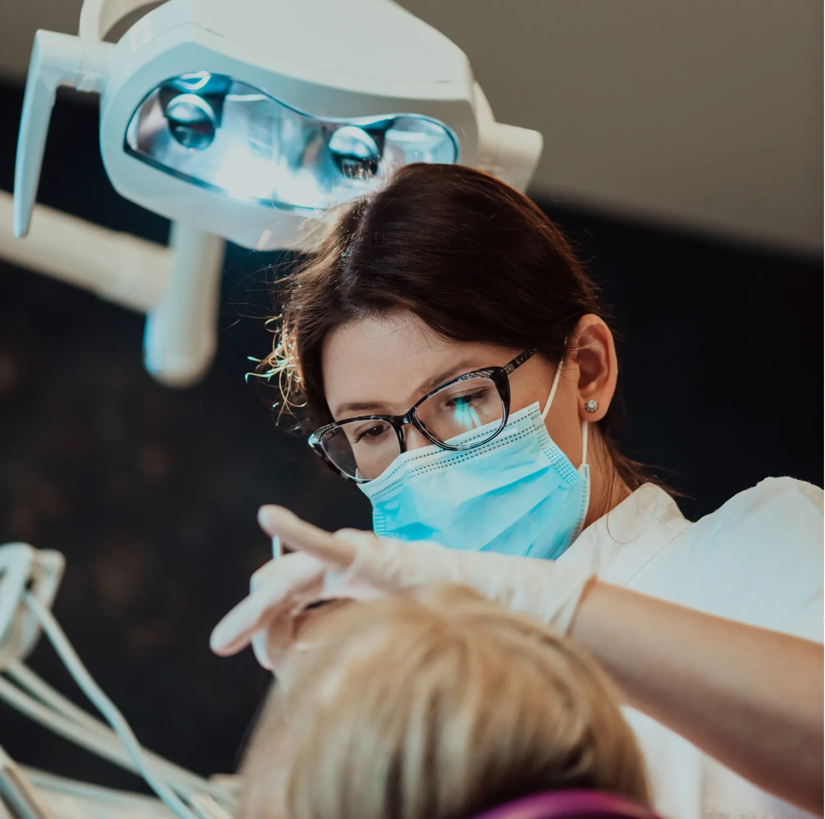 Dentist wearing glasses and a mask examining a patient under a dental light.
