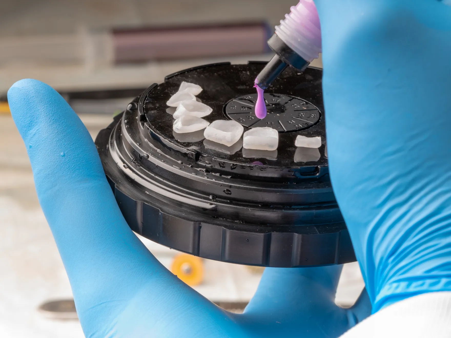 Gloved hand holding a round black tray with dental veneers while a dental tool dispenses purple liquid onto one veneer.