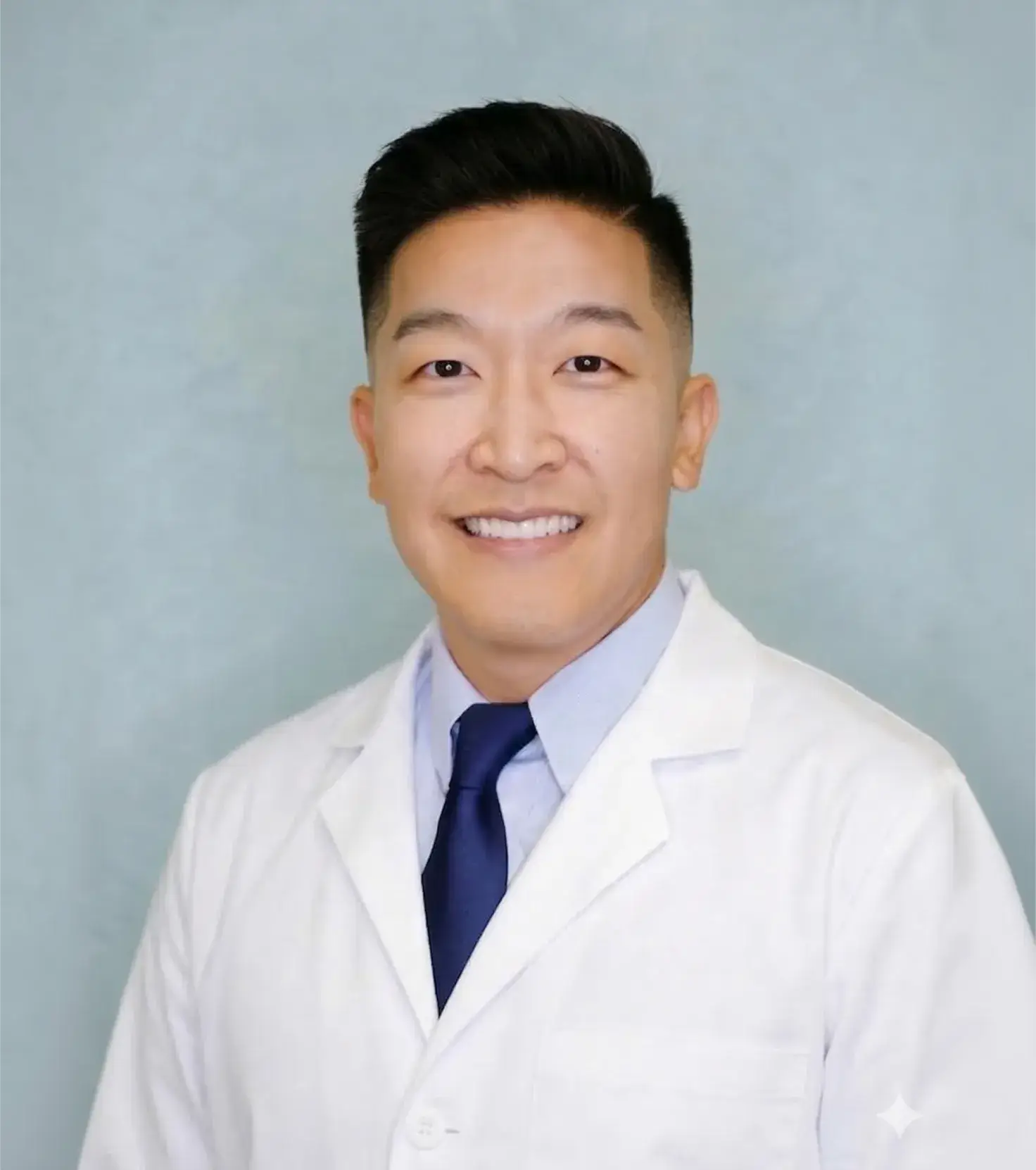 Smiling male doctor with black hair wearing a white coat, blue shirt, and dark blue tie against a light blue background.