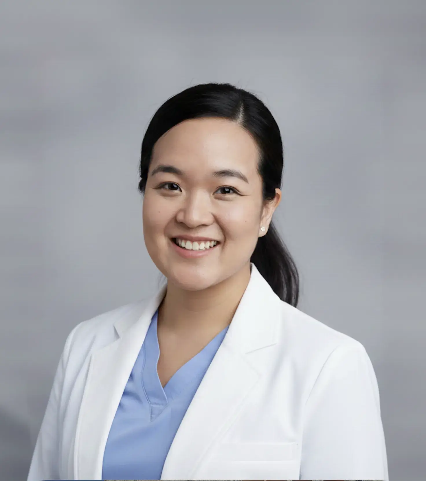 Smiling female doctor wearing a white coat and light blue scrubs against a gray background.