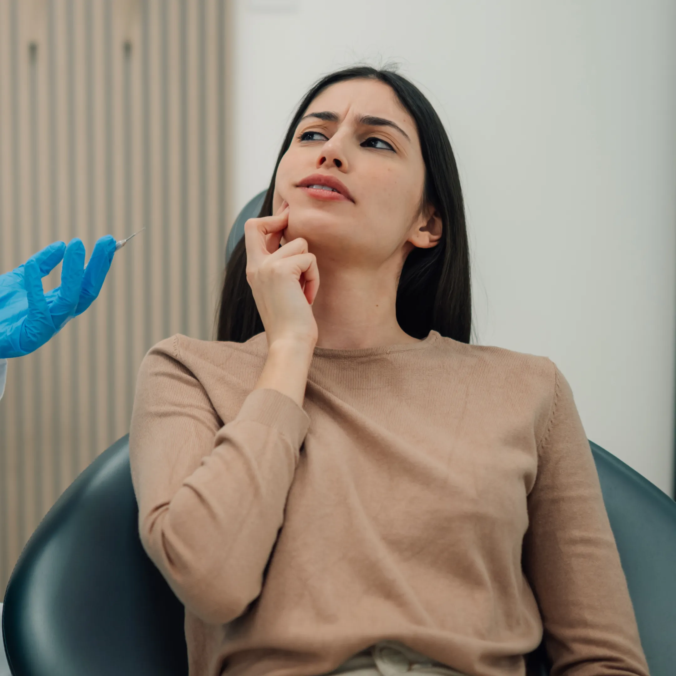 Dentist wearing gloves and a mask talking to a woman in a dental chair who is touching her cheek in discomfort.