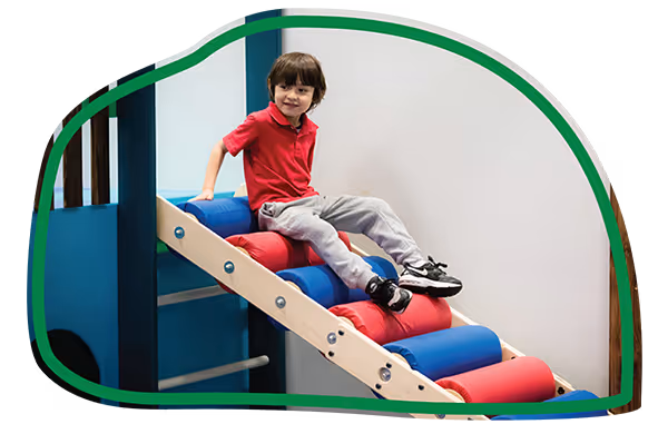 Young boy in a red shirt sitting and sliding down a colorful indoor playground slide.