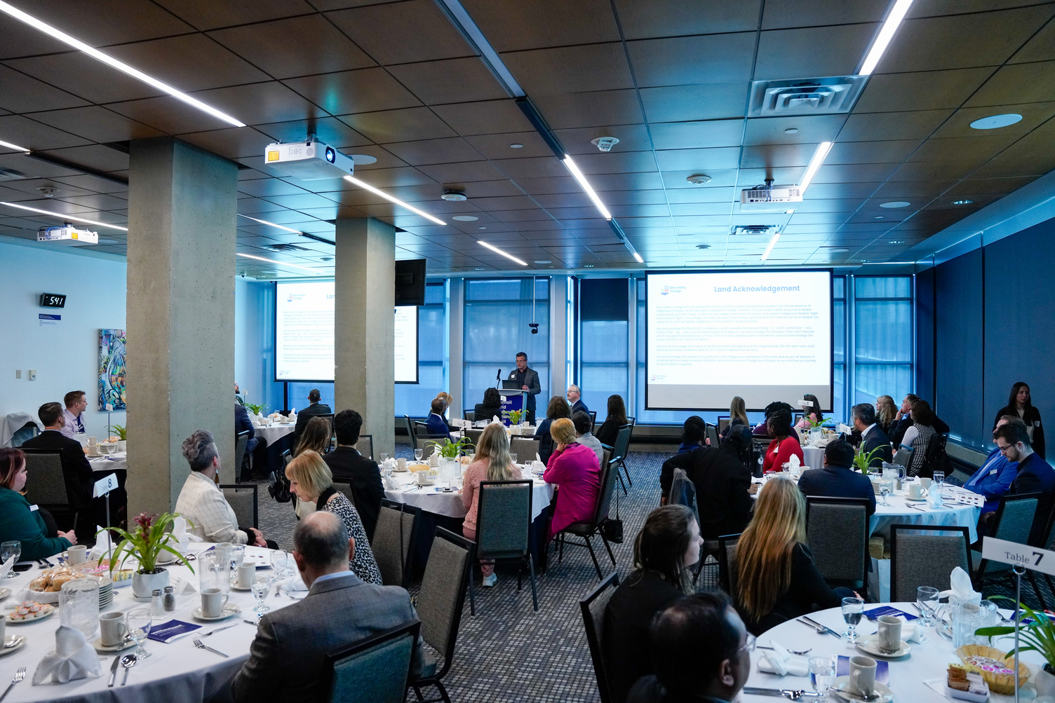 The 2025 VentureQuest event taking place at Bow Valley College in the Esmail and Safana Bharwani Hall. The hall is filled with people sitting at tables and a speaker at the front welcoming guests.