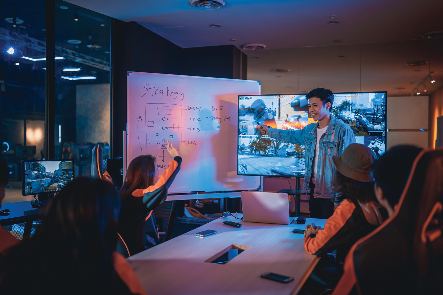 A group of young people in a gaming room sit around a table while one person stands at a whiteboard explaining a strategy with diagrams. Monitors display a video game, and the room is lit with blue and orange lights.