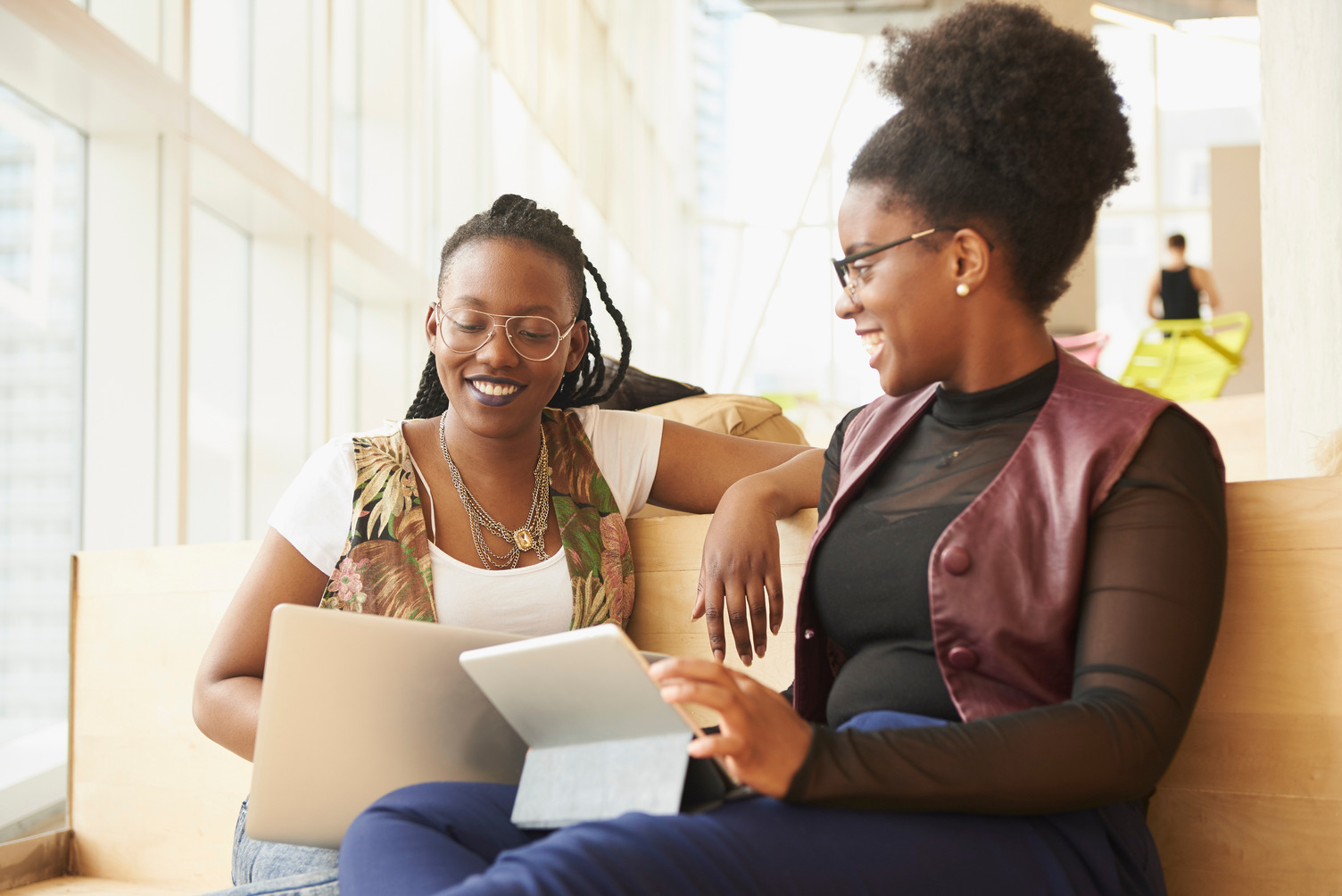 Two women sit on a bench in a bright, modern space, smiling and chatting while using a laptop and a tablet. One wears glasses and braids; the other has an afro and wears glasses.