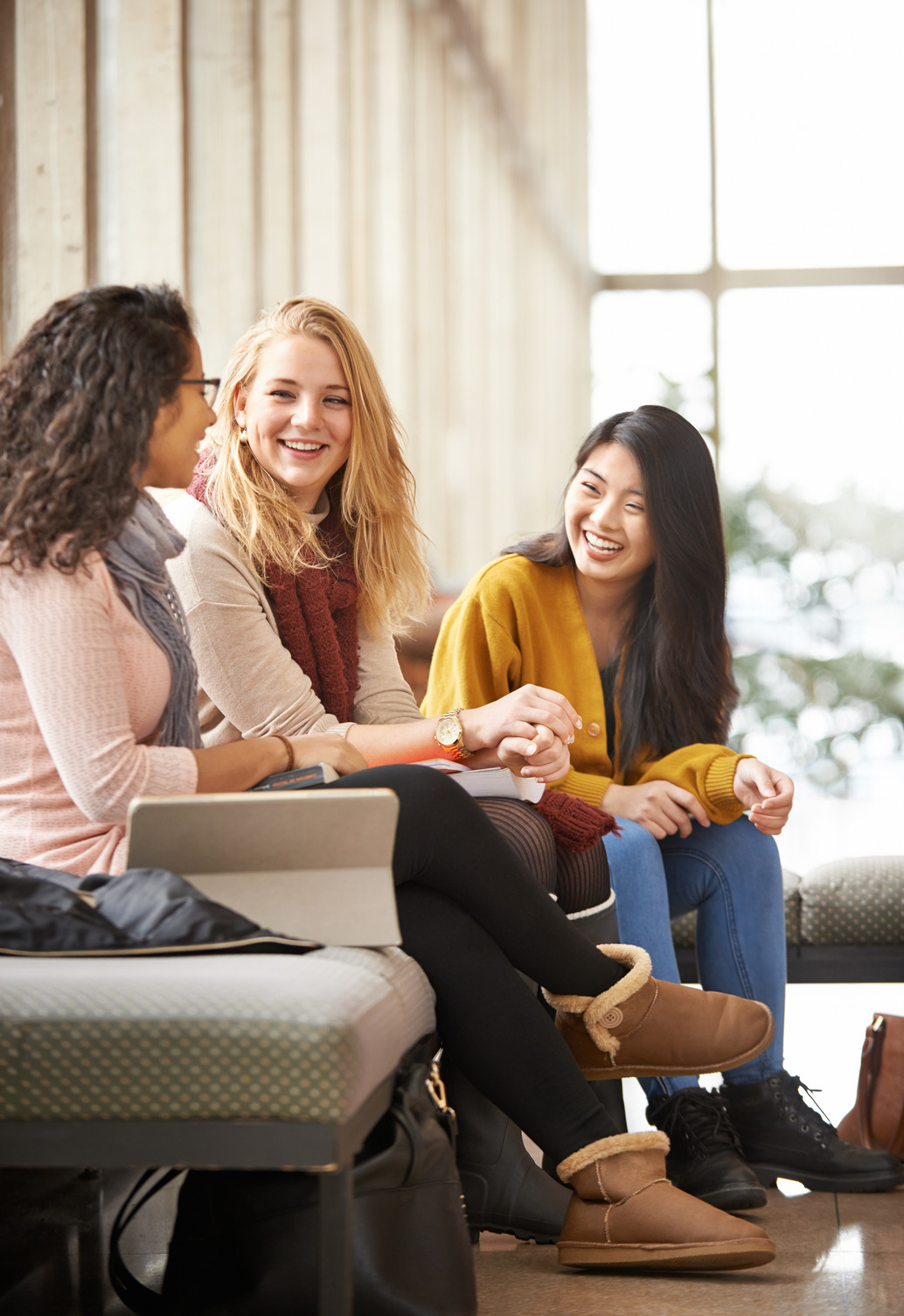 Three young women who are students sitting together indoors on a bench, talking and laughing. They have notebooks and bags beside them, enjoying a friendly conversation in a bright, sunlit space.