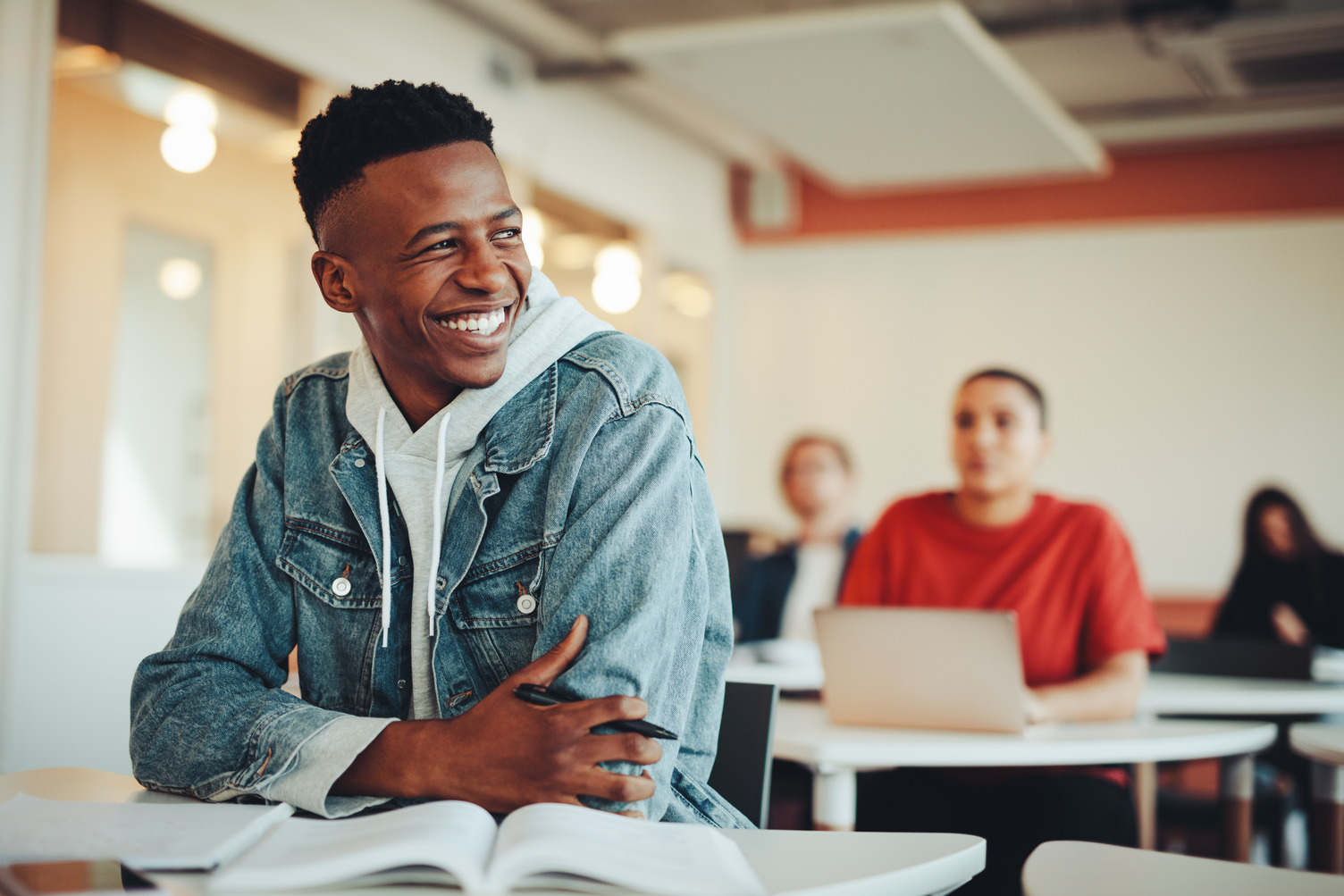 Young man in a denim jacket smiles while sitting at a desk with an open book in a bright classroom; other students are visible in the background.