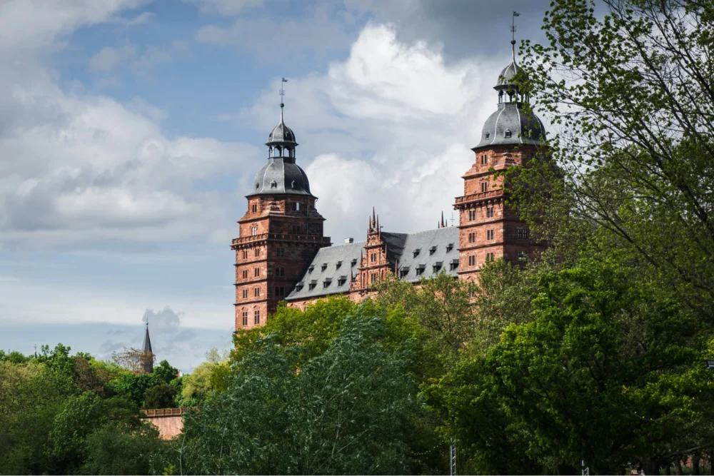 Historic castle with two tall towers and a steep roof surrounded by green trees under a cloudy sky.