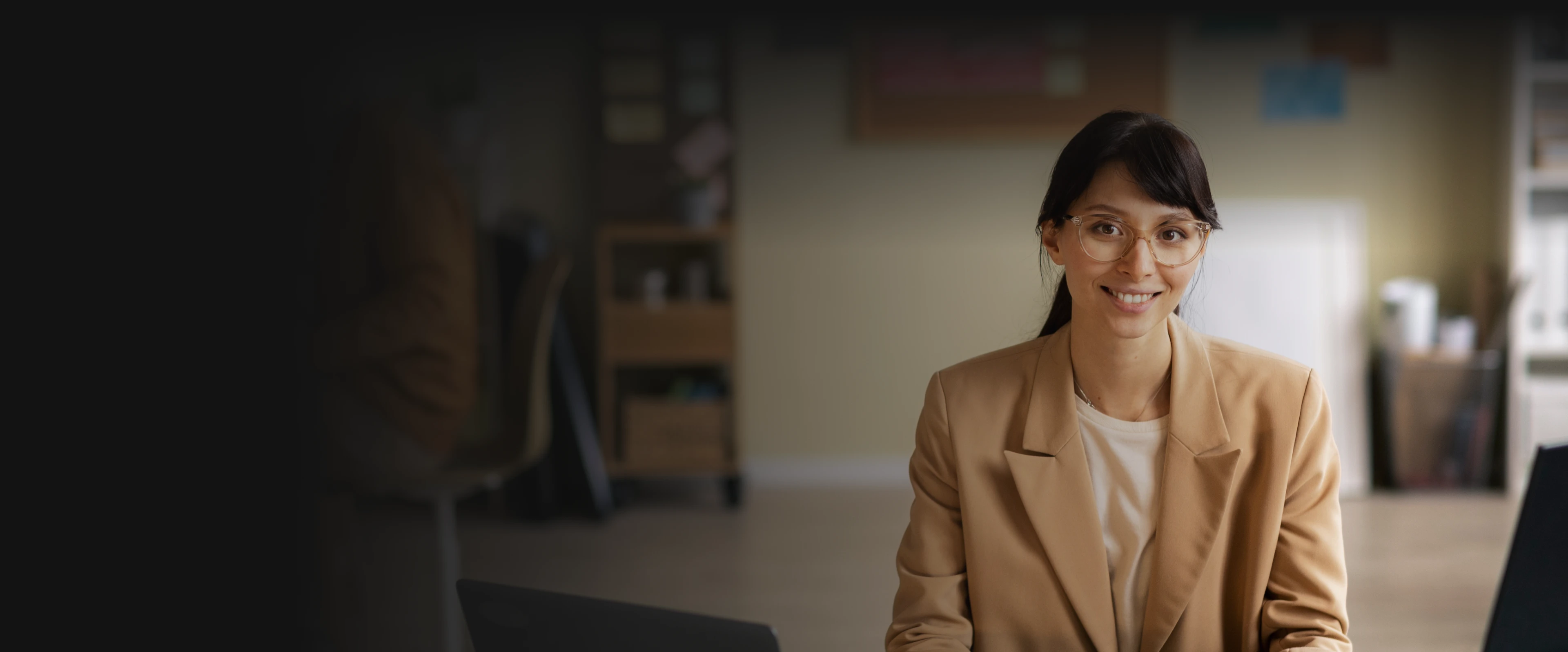 Smiling woman in glasses and beige blazer sitting in an office environment with blurred background.
