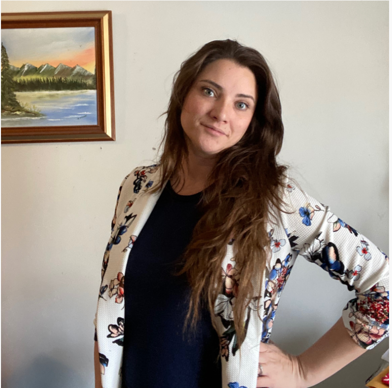 Woman with long brown hair wearing a floral blazer over a black top, standing with one hand on her hip beside a framed landscape painting.