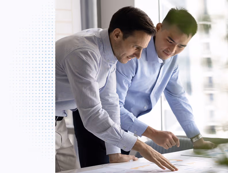 Two men in dress shirts leaning over a desk reviewing papers with charts and graphs.