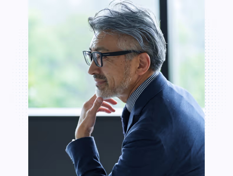 Middle-aged man with gray hair and glasses, in a suit, thoughtfully touching his chin indoors.