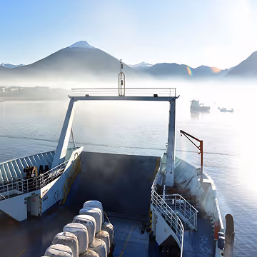 View from a ferry deck with mountains, mist, and boats on calm water in the distance.