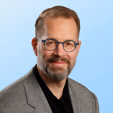 Man with short brown hair, beard, glasses wearing a grey herringbone jacket and black shirt against blue background.