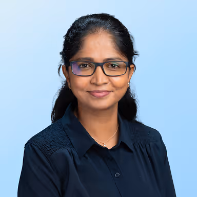 Smiling woman with glasses and long dark hair wearing a dark shirt against a light blue background.