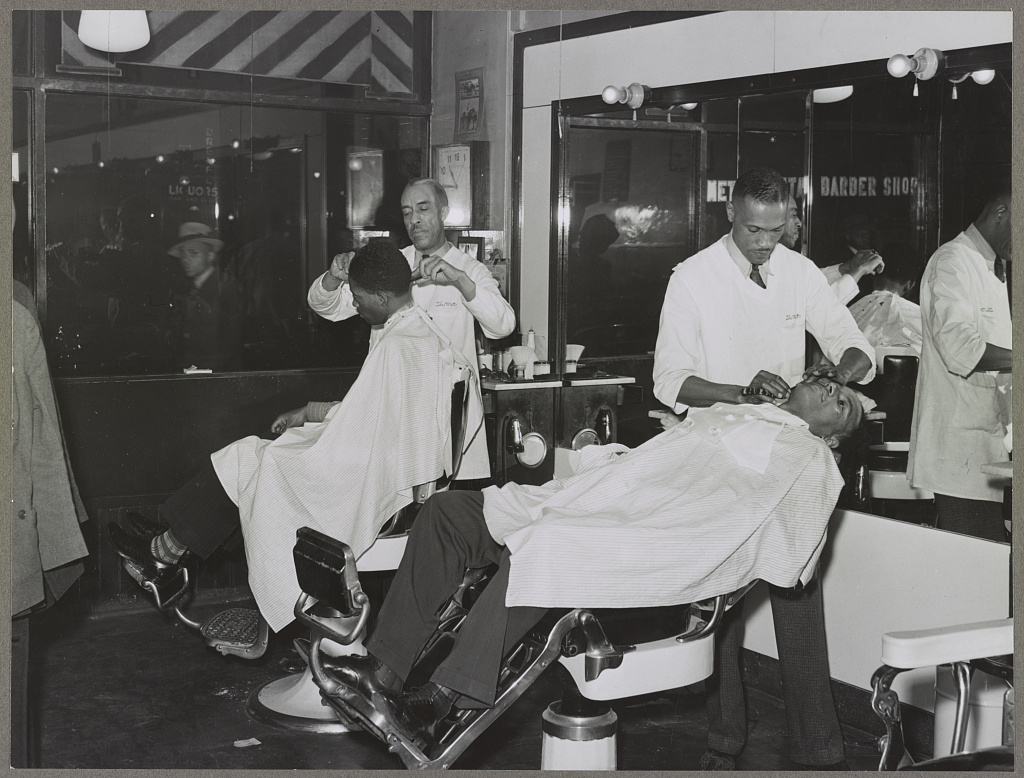 Black-and-white photograph of men receiving haircuts and shaves inside a mid-century barbershop.