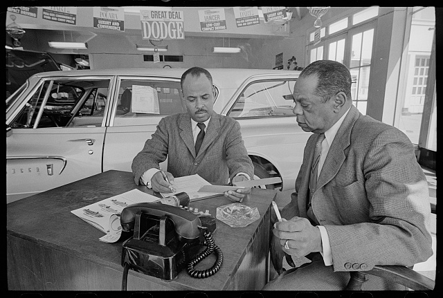 Black-and-white photograph of two men in suits seated at a desk inside a car dealership.